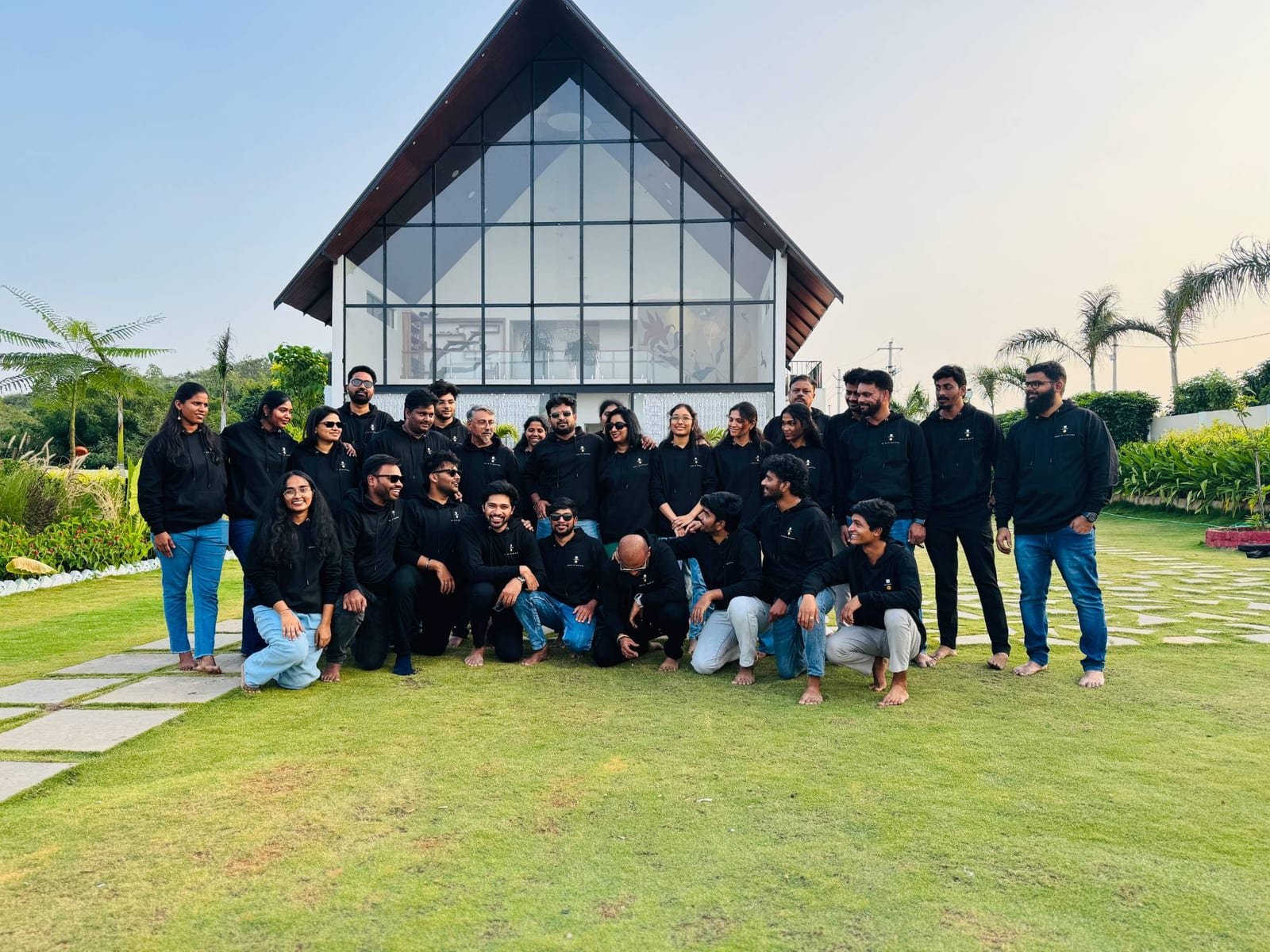 The House of Investors team, about 30 people in matching black hoodies, standing and kneeling on a lawn in front of a tall glass house.